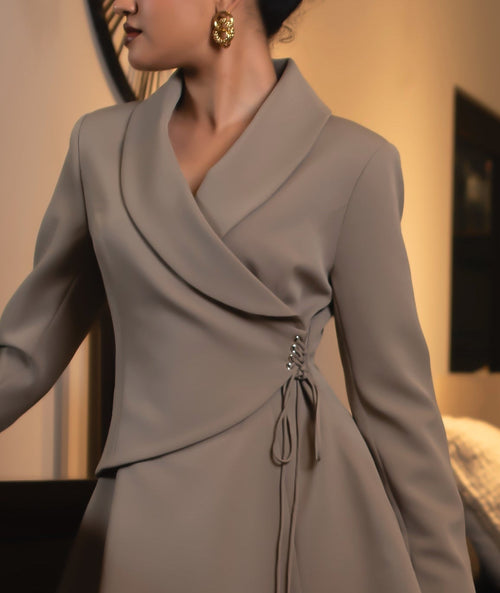 A woman models a taupe wrap-style mini dress with structured shoulders and a tie waist, standing in a dimly lit room next to a dark wooden dresser.