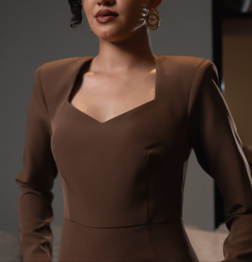 A woman wearing a fitted brown long-sleeve dress with structured shoulders and a square neckline, standing in a softly lit indoor setting with a sofa and shelving in the background.